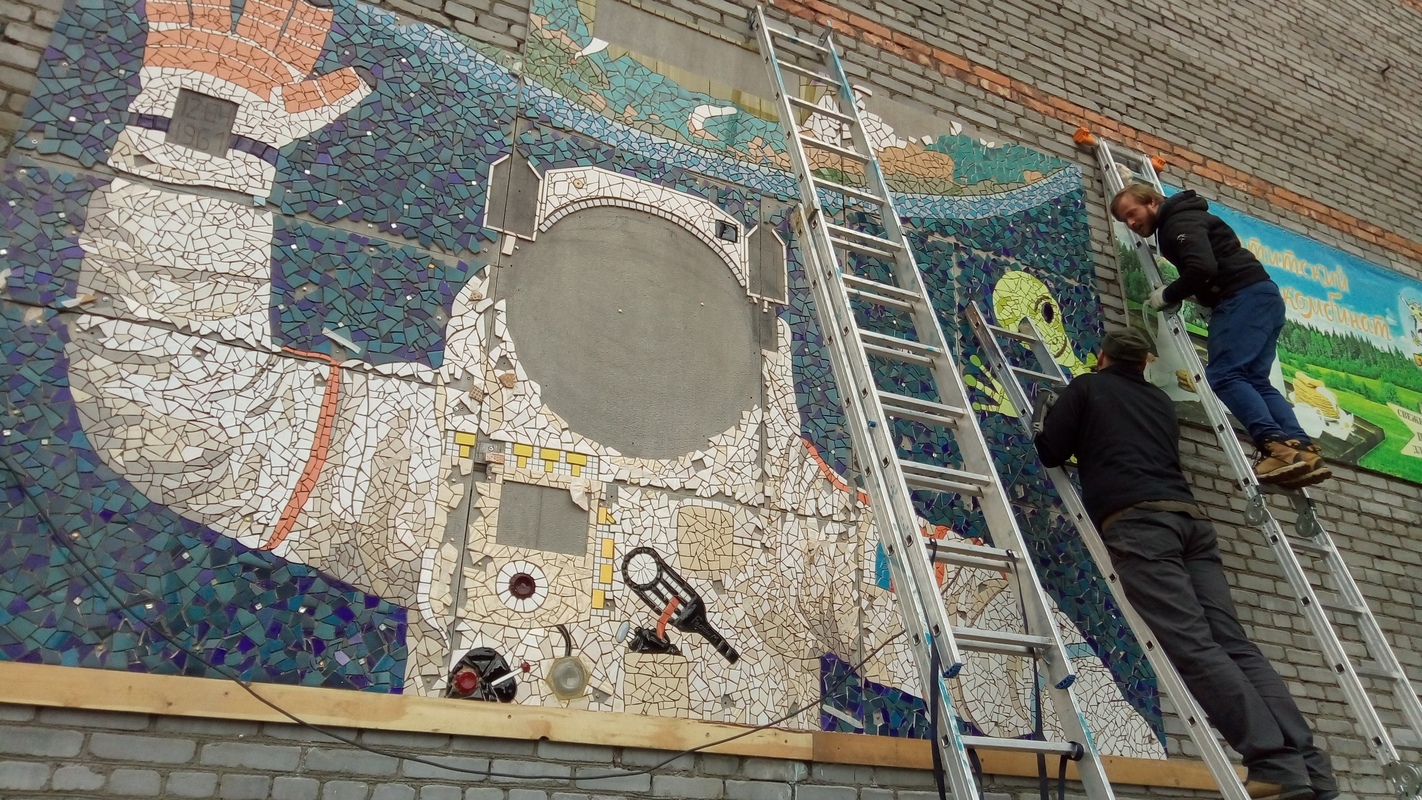 Two people on ladders installing a colourful cosmonaut mosaic on a brick wall in Apatity